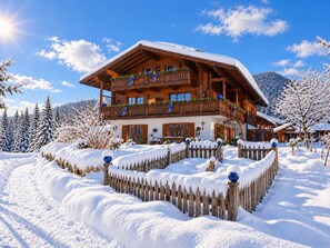 A large wooden chalet with multiple balconies, surrounded by snow-covered trees and a wooden fence.