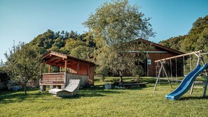 En legeplads med rutsjebane og gyngestole, et træpavillon og en bygning i et græsareal med træer.