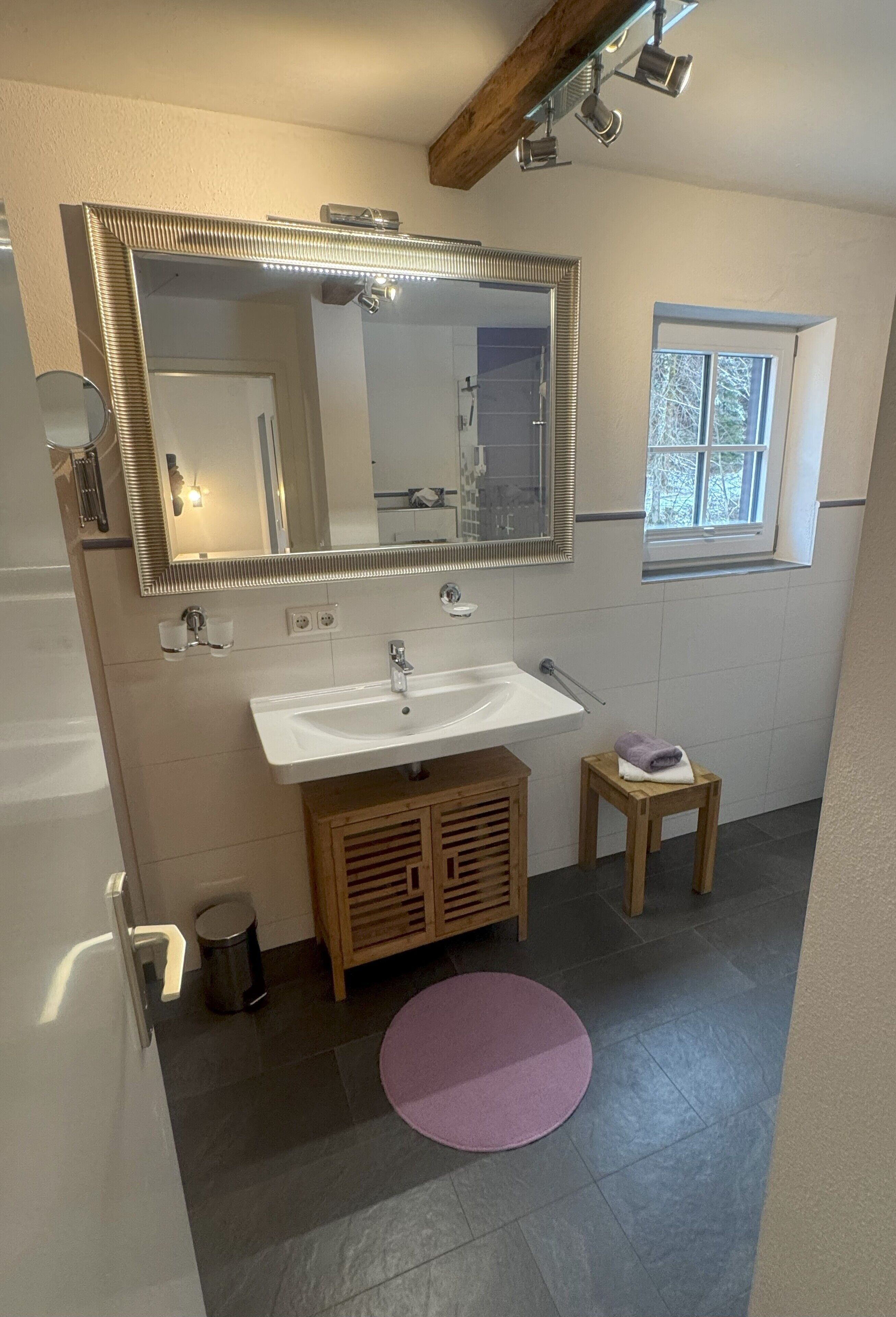A bathroom with a large mirror, a sink, a wooden cabinet, a small stool, a trash can, and a window.