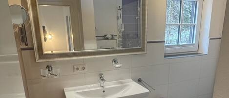A bathroom with a large mirror, a sink, a wooden cabinet, a small stool, a trash can, and a window.