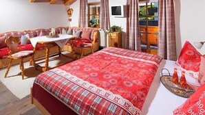 A cozy room with a wooden ceiling, a bed with a red and white checkered bedspread, a wooden bench with red cushions, a table with a white tablecloth, and a window with curtains. - Ferienwohnung Watzmann für 2-7 Personen, ca. 90 m², 2 sep. Schlafzimmer, Wohnküche, Balkon, 2 Zustellbetten (Bischofswiesen)
