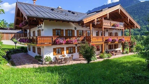 A two-story chalet with a wooden balcony, a small garden, and a mountain backdrop. - Ferienwohnung Watzmann für 2-7 Personen, ca. 90 m², 2 sep. Schlafzimmer, Wohnküche, Balkon, 2 Zustellbetten (Bischofswiesen)