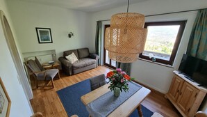 Een gezellige woonkamer met een bank, een houten tafel met een bloemenvaas, een televisie en een groot raam met gordijnen.