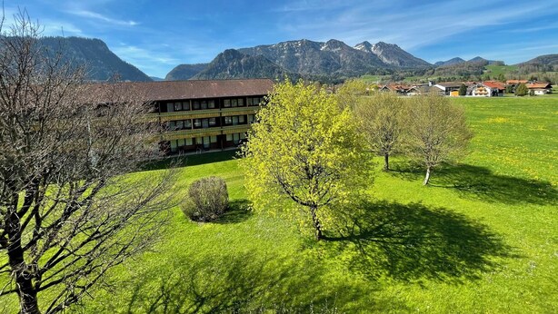 A building with a red-tiled roof and multiple windows, surrounded by green fields and mountains in the background. - Vacation apartment C, 4 pers. with balcony or terrace, bedroom, 40 to 45 sqm (Inzell)