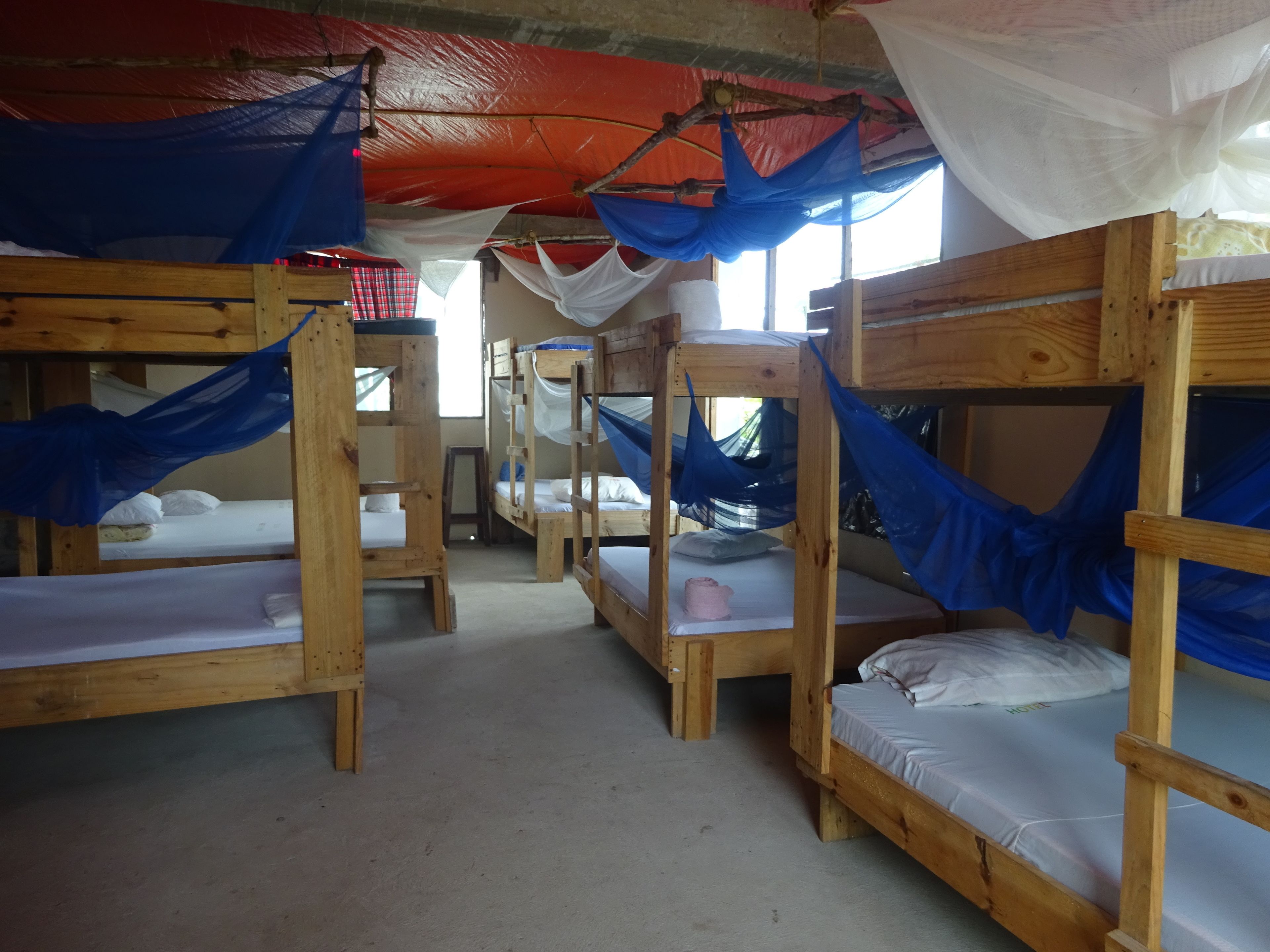 A row of wooden bunk beds with mosquito netting, a red tarp overhead, and a window providing natural light.
