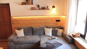 Une pièce chaleureuse avec un canapé d’angle gris, une tête de lit en bois, une petite étagère avec des livres et des figurines, et une fenêtre donnant sur un beau paysage.