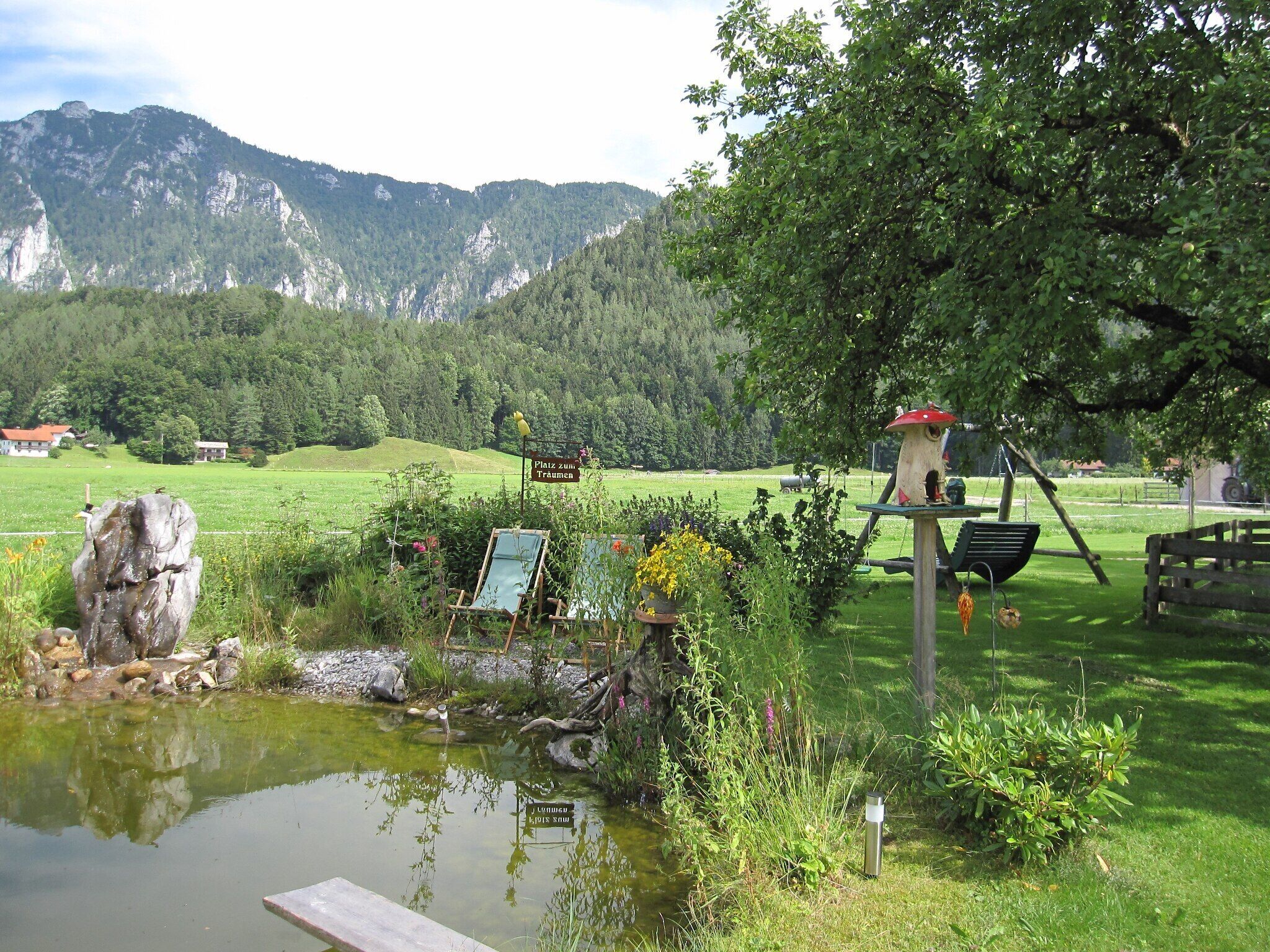 Ruhige Seelandschaft mit Holzsteg, Vogelhaus und einem Schild in einer natürlichen Umgebung, mit Bergen im Hintergrund.