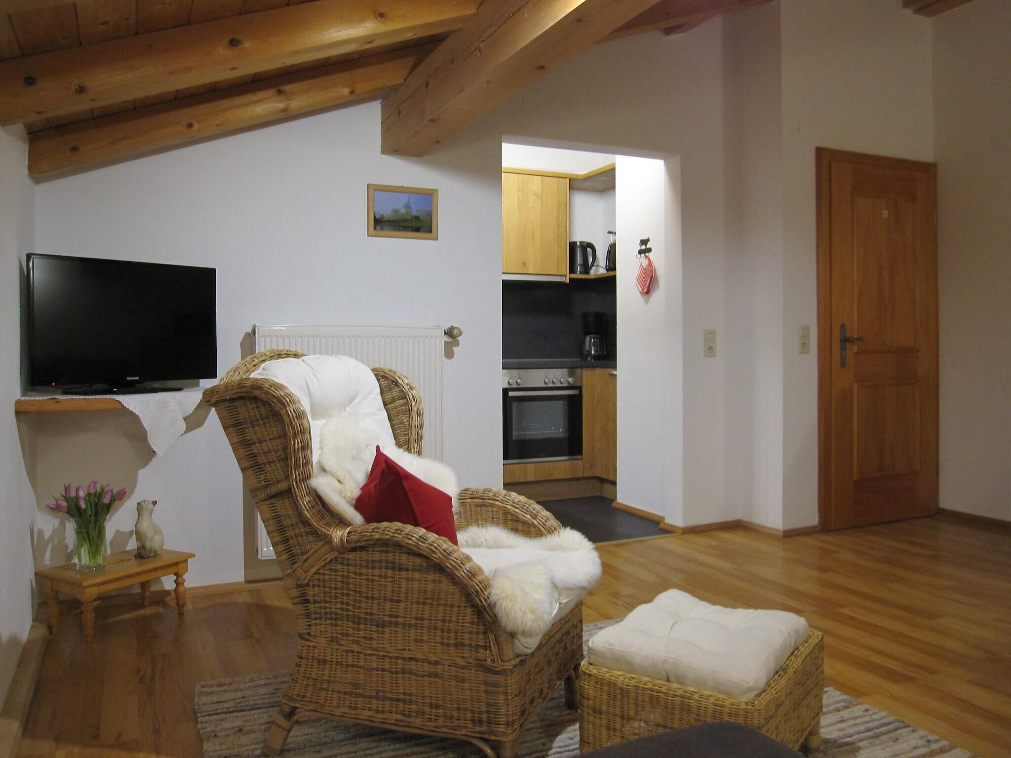 Een woonkamer met een rieten fauteuil, een televisie, een klein tafeltje met een vaas, en een keuken op de achtergrond.