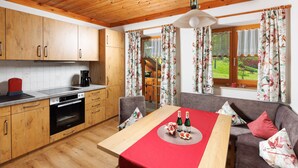 Une cuisine avec des meubles de rangement en bois, une table à manger recouverte d’une nappe rouge et un canapé agrémenté de coussins à motifs floraux.