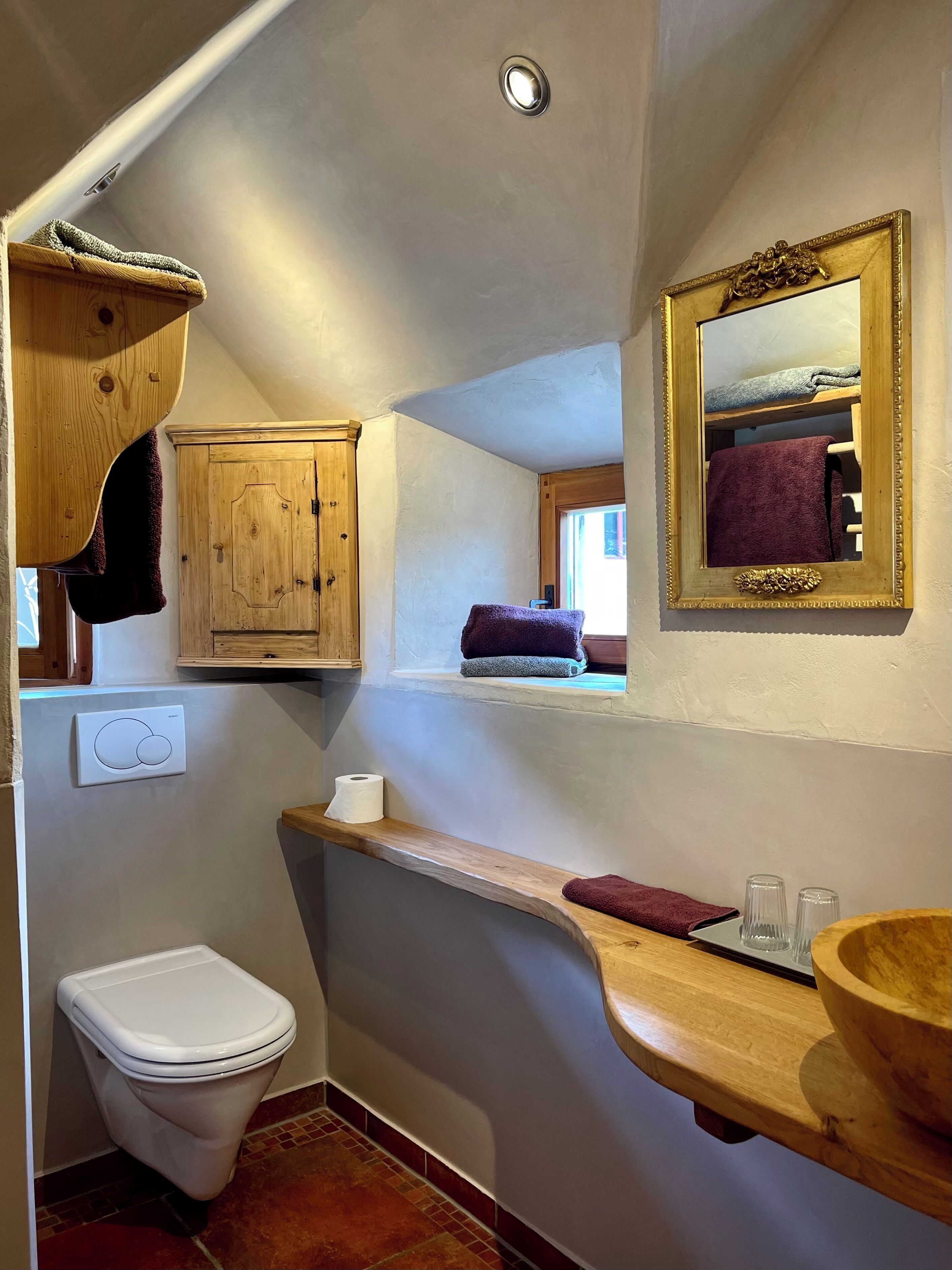 Een kleine badkamer met een houten wastafelmeubel, een toilet, een spiegel met gouden lijst en een handdoekhouder.
