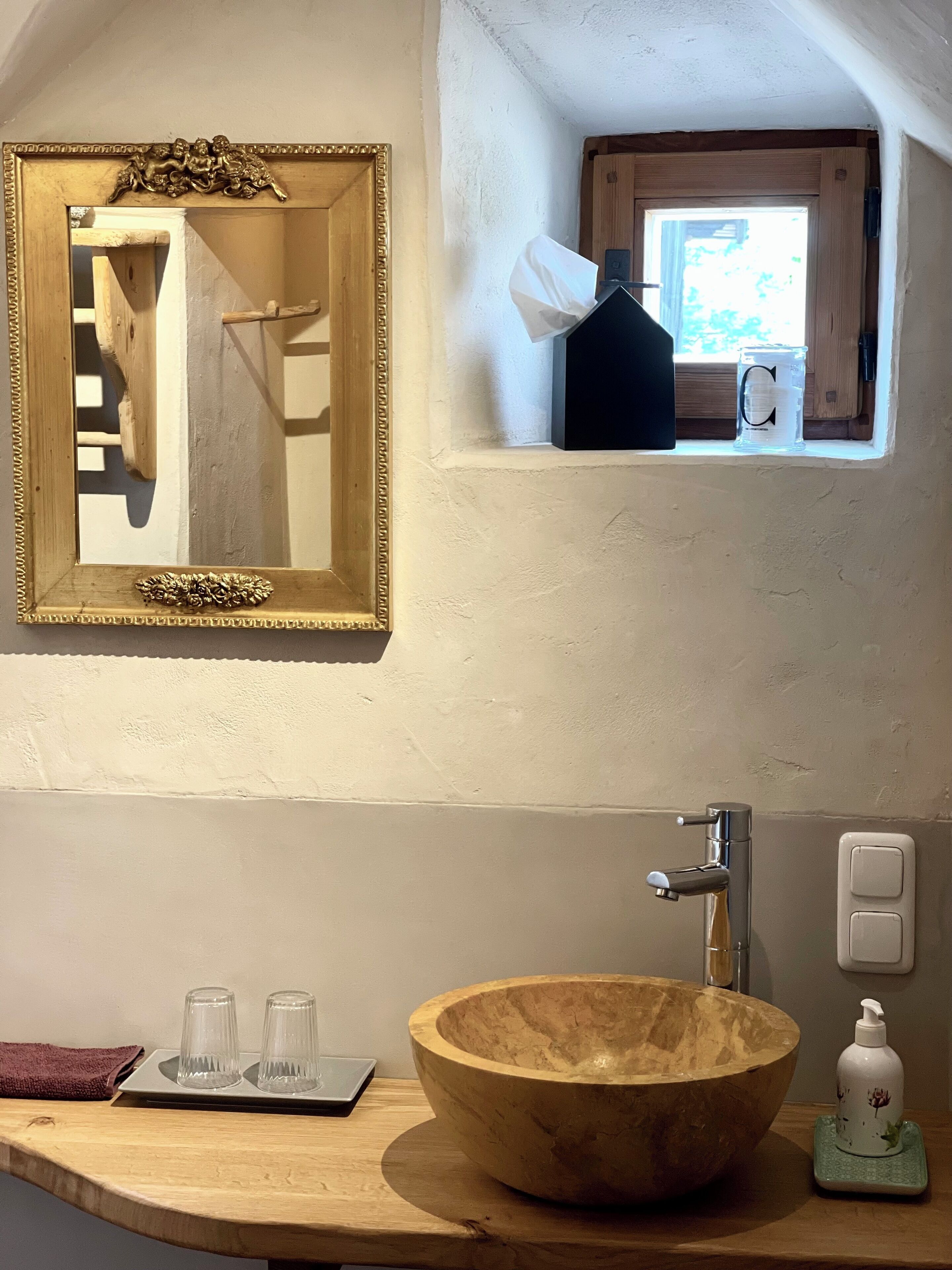 Ein Badezimmer mit einem Holzwaschbecken, einem Spiegel mit aufwendiger Rahmung und einem kleinen Fenster, auf dem eine Kerze und eine Taschentuchbox stehen.