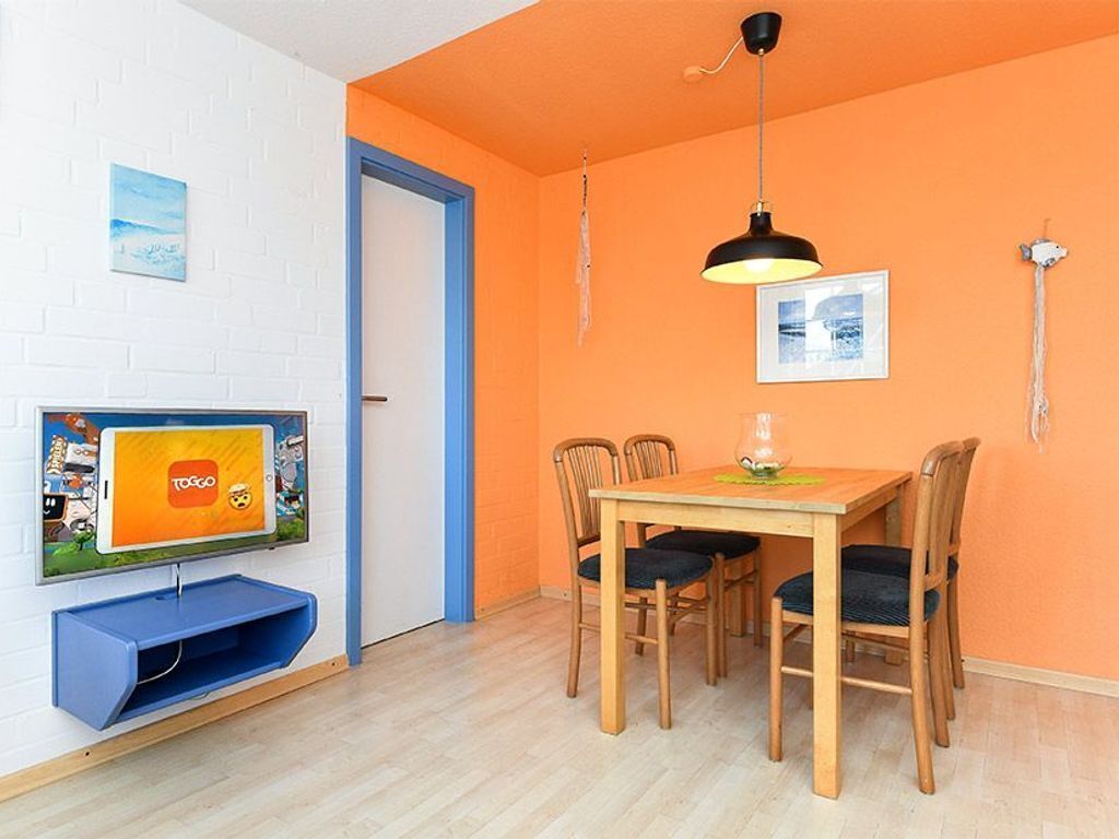 Ein Zimmer mit einem Holztisch, vier Stühlen, einem an der Wand montierten Fernseher und einer Tür mit blauer Umrandung.
