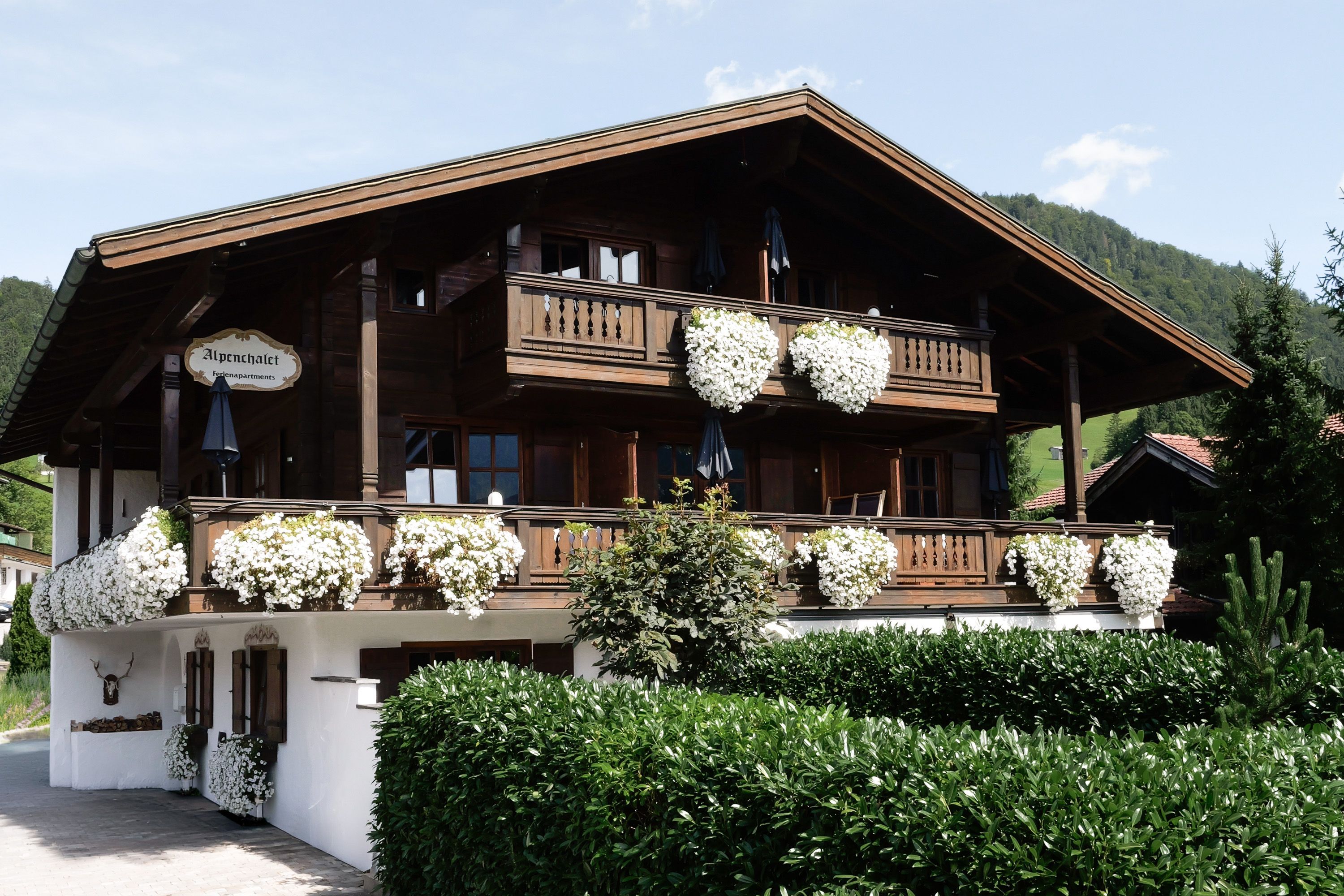 A wooden chalet with a balcony adorned with white flowers, a sign indicating it's a hotel, and a mountainous backdrop.