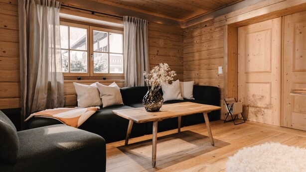 A cozy cabin interior with a wooden floor, a sofa, a table with a vase of flowers, and a window with curtains. - (8) Vier-Raum-Ferienwohnung 85qm, 2 Bäder, 3 Schlafzimmer, Küche und Balkon (Reit im Winkl)
