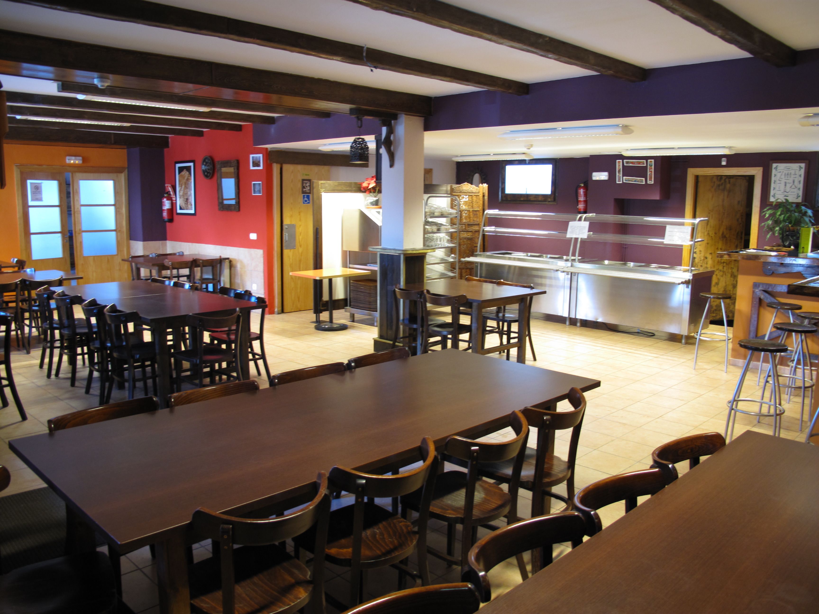 A restaurant with multiple wooden tables and chairs, a buffet area with a stainless steel counter, and a red wall with framed pictures.