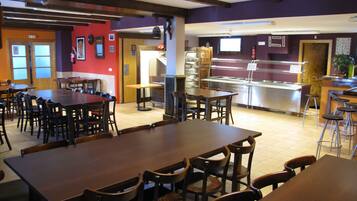 A restaurant with multiple wooden tables and chairs, a buffet area with a stainless steel counter, and a red wall with framed pictures.