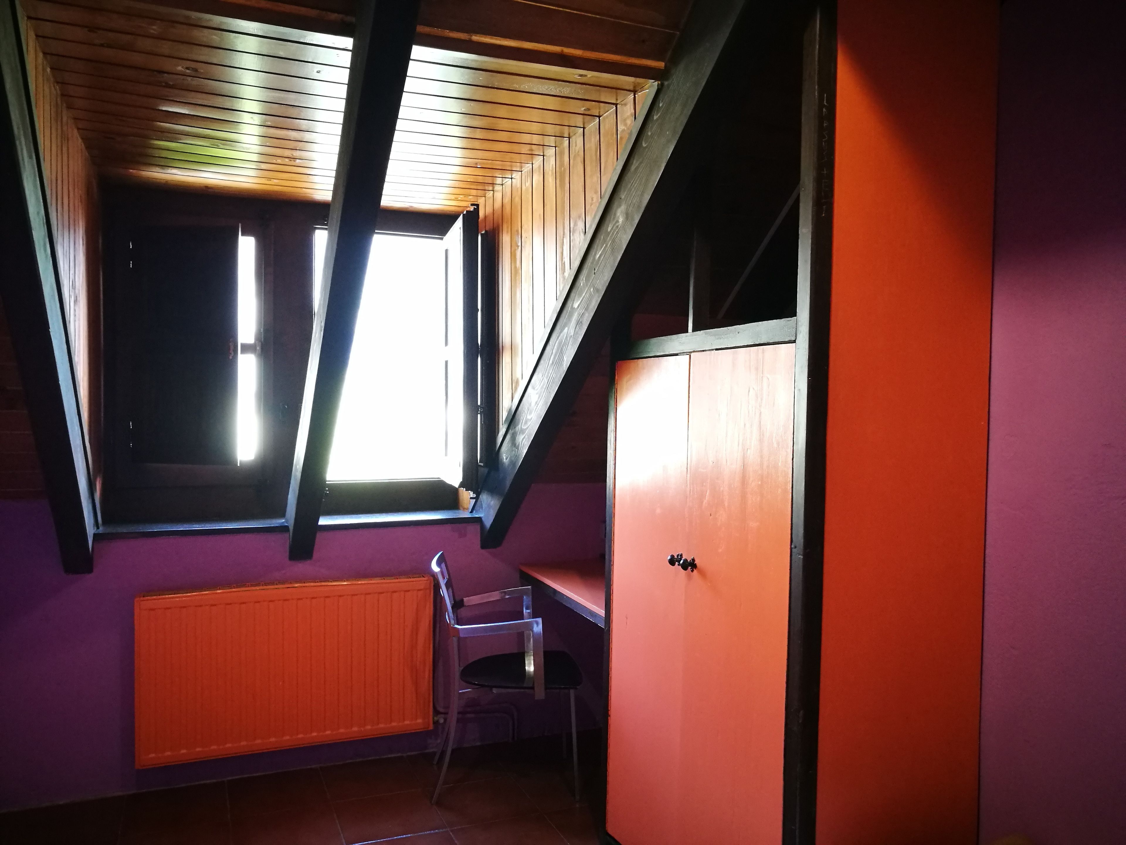 Una habitación con techo inclinado, una ventana, un radiador y una silla.