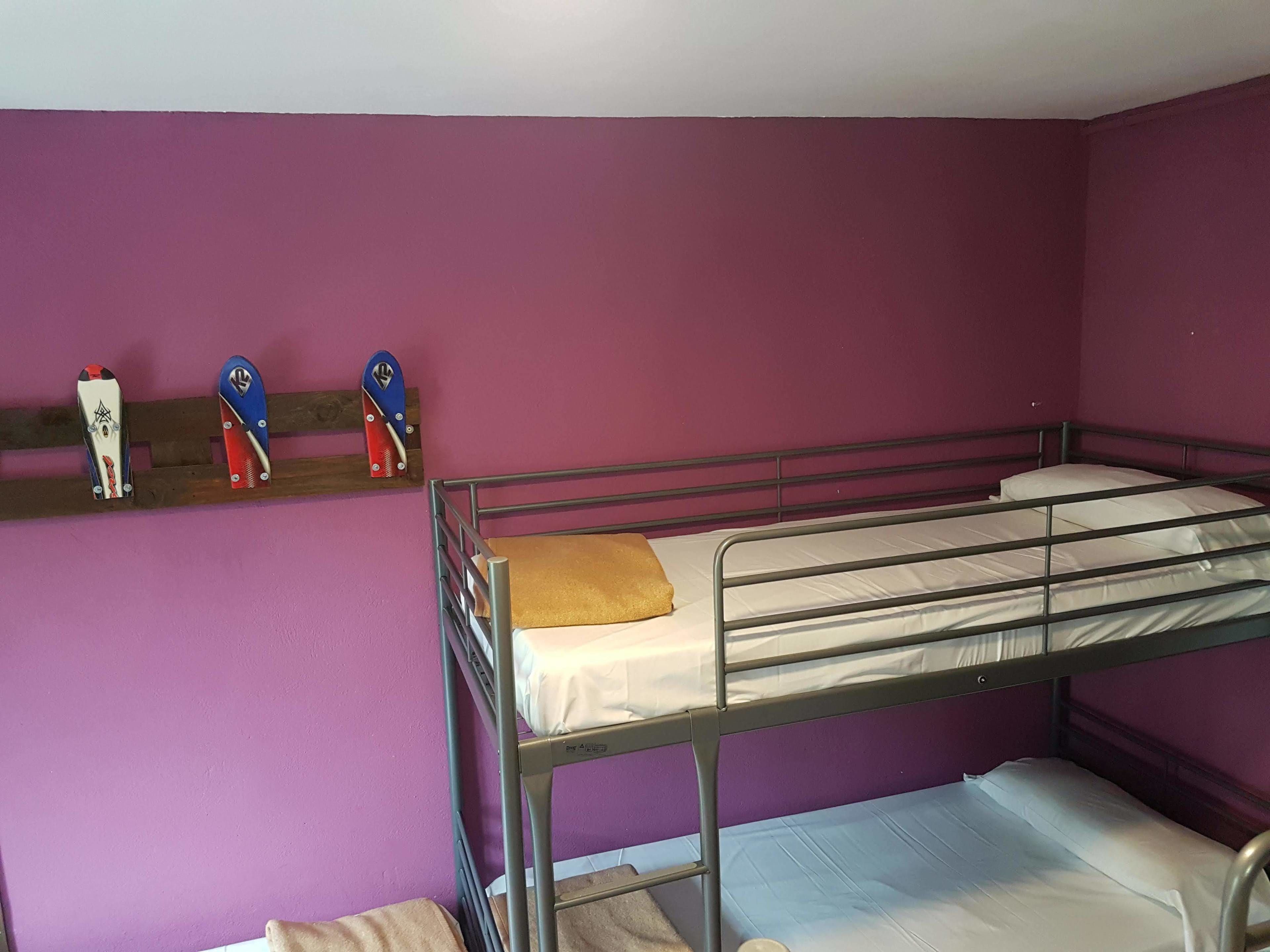 Quarto com beliches, paredes roxas, uma prateleira de madeira com facas e estrutura de cama de metal.