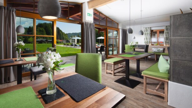 A modern hotel lobby with wooden flooring, green cushioned seating, and large windows offering a view of a green landscape.