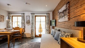 A room with a wooden wall, a sofa, a TV, a dining table with chairs, and a window with curtains.