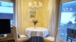 Zona pranzo con tavolo rotondo, due sedie, un lampadario, un vaso con fiori e vista su un paesaggio innevato attraverso un'ampia finestra.