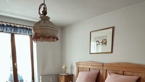 A wooden bed with pink bedding, a bedside table with a lamp, and a window with curtains. - Ferienwohnung 60 m², 1-2 Personen, Schlafzimmer, Kabel-TV, Balkon, Nichtraucher (Unterwössen)