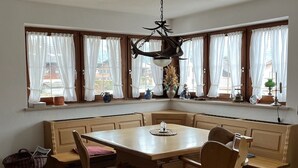 A dining area with a wooden table and benches, large windows with curtains, and a hanging light fixture. - Ferienwohnung 60 m², 1-2 Personen, Schlafzimmer, Kabel-TV, Balkon, Nichtraucher (Unterwössen)