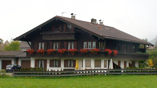 A two-story wooden house with a balcony, a car parked in front, and a wooden fence. - Ferienwohnung 60 m², 1-2 Personen, Schlafzimmer, Kabel-TV, Balkon, Nichtraucher (Unterwössen)