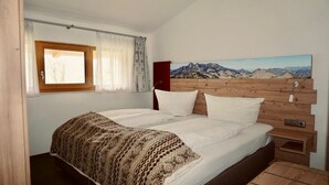 A bedroom with a bed, a window with curtains, a wooden headboard, and a painting of mountains. - Vacation apartment 60sqm (Brannenburg)