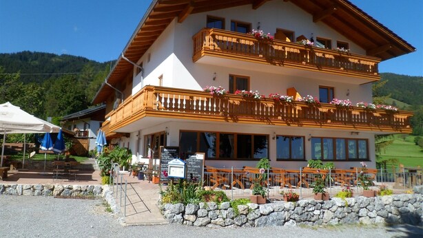 A two-story building with balconies adorned with flowers, a stone wall in front, and a signboard. - Vacation apartment 60sqm (Brannenburg)