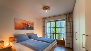 A bedroom with two beds, a large window with curtains, a painting on the wall, and a ceiling light fixture. - Apartment Unternberg 58sqm balcony, kitchen, 1 bedroom, 1 bed-sitting room, 2 p. (Ruhpolding)