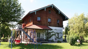 Una casa di legno con tetto rosso, un’area giochi con scivolo e altalene, e un giardino recintato da una staccionata di legno.