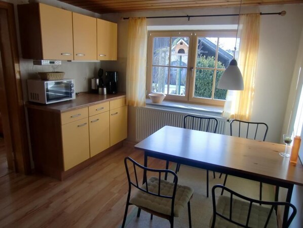 Cocina compacta con horno integrado, mesa de comedor con cuatro sillas y ventana con vista a una casa y follaje.