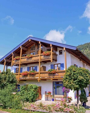 A two-story wooden building with balconies, a front door, and a garden with flowers. - Ein-Raum-Ferienwohnung (4) 26qm, Dusche/WC, Küchenzeile, Balkon (Reit im Winkl)