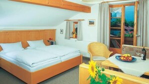 Um quarto com uma cama, uma cadeira, uma mesa com uma travessa de fruta, um vaso com um girassol, uma janela com cortinas e um lustre.