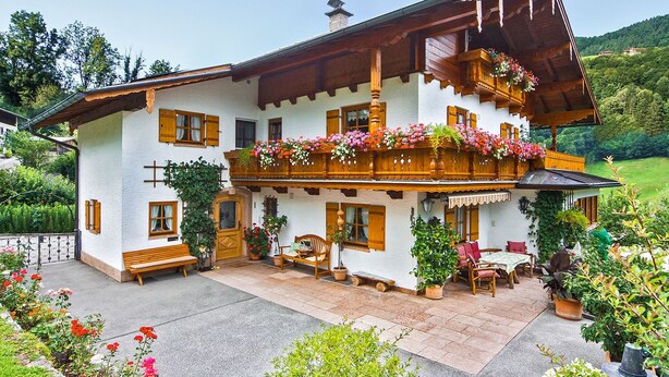 A two-story chalet with a balcony adorned with flowers, a wooden bench, and a patio area with outdoor seating. - Ferienwohnung für 1-2 Personen, ca. 45qm (Marktschellenberg)