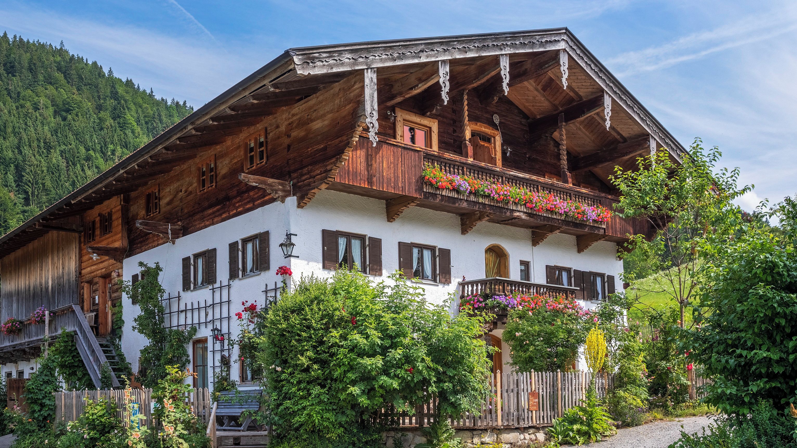 Un chalet traditionnel en bois, doté d’un balcon, entouré d’une végétation luxuriante et bordé par une forêt.