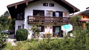 A two-story building with a balcony, a car parked in front, and a garden with trees and shrubs. - Apartment 82sqm, terrace, kitchen, 2 bedrooms, 1 bed-sitting room, WLAN, 6 p. (Ruhpolding)