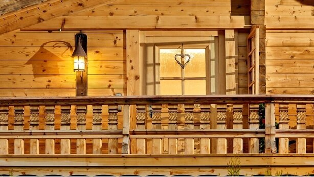 Una cabaña de madera con un balcón, una ventana decorada con forma de corazón y una lámpara fijada a la pared.
