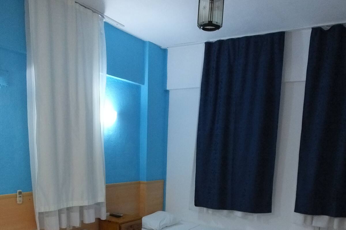 Une chambre d’hôtel avec trois lits simples, une tête de lit en bois, une table de chevet et une grande fenêtre avec des rideaux.