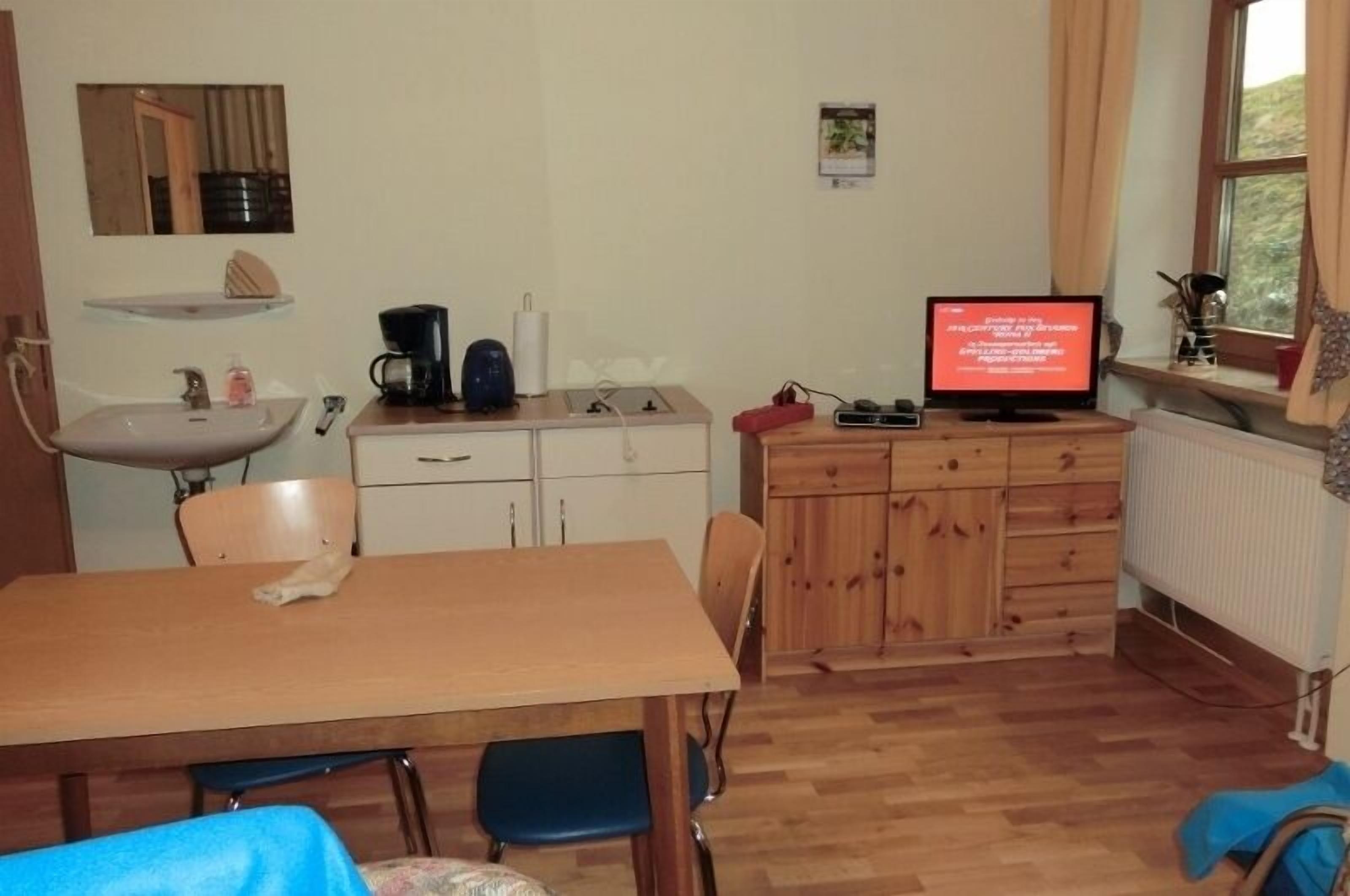 Une pièce comprenant une table en bois, des chaises, un petit lavabo, un meuble et un téléviseur affichant un message.