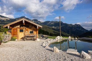 Una cabaña de madera con terraza de madera, una piscina pequeña y montañas al fondo.