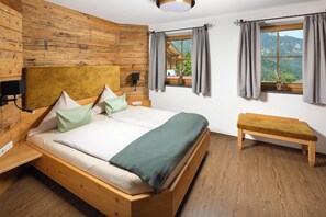 Una cama de madera con un banco, una ventana con cortinas y vistas a las montañas.