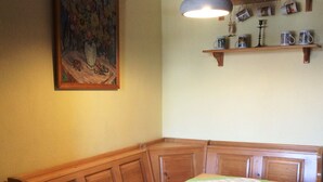 Een kamer met een houten bank, een tafel met een geruit tafelkleed, een hanglamp en een plank met mokken en een kaars.