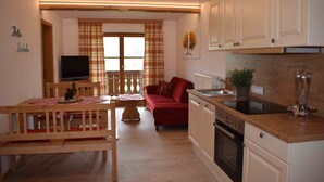 A compact living space with a dining area, a sofa, a TV, a kitchen with a stove, and a window with curtains. - Apartment Rauschberg 56 sqm, balcony, kitchenette, 2 bedrooms, WLAN, 4 pers (Ruhpolding)