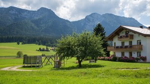 A two-story building with a balcony, surrounded by a grassy area with a swing set and a tree. Mountains are visible in the background. - Apartment Rauschberg 56 sqm, balcony, kitchenette, 2 bedrooms, WLAN, 4 pers (Ruhpolding)