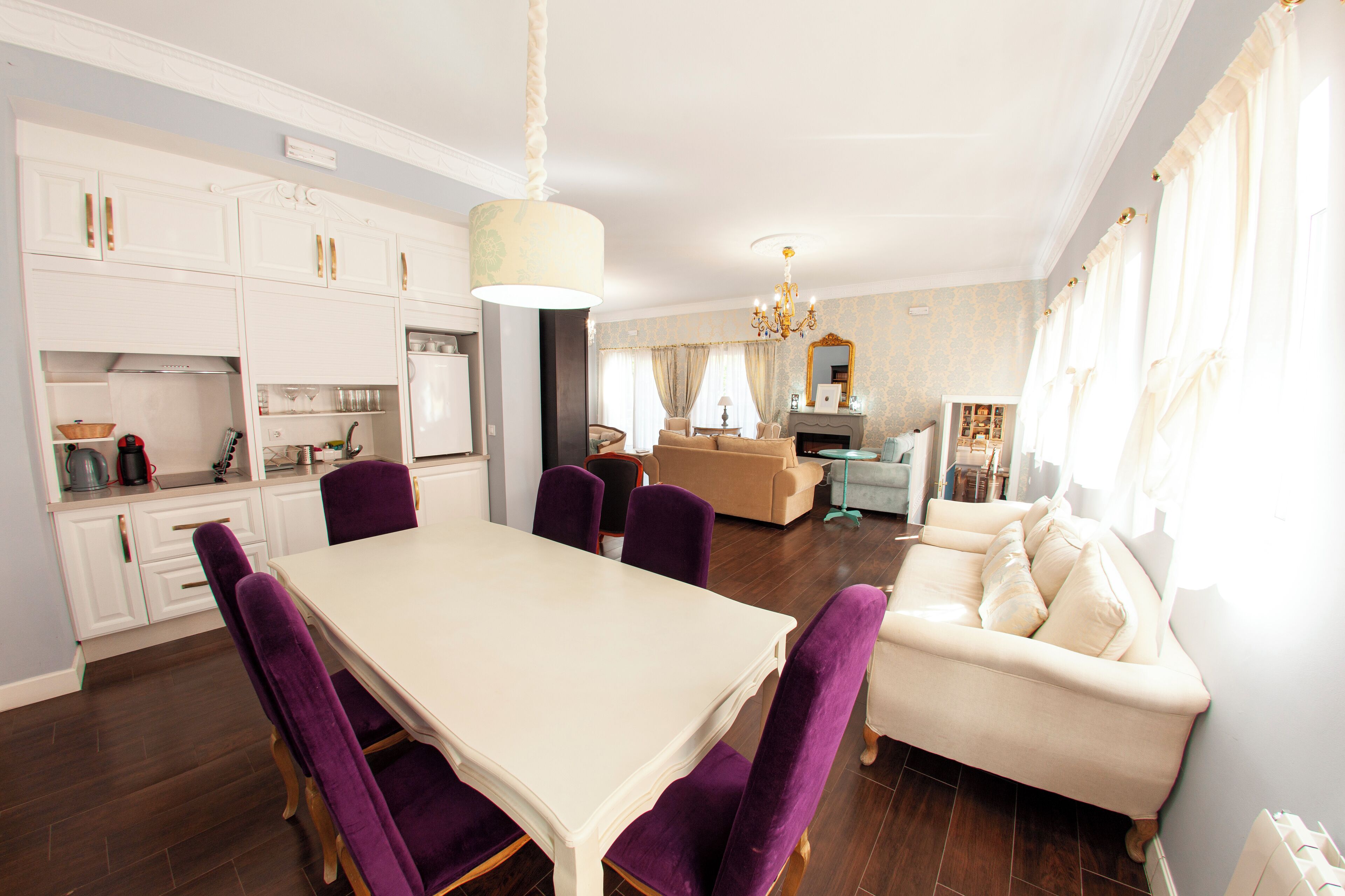 A spacious living area with a dining table, purple chairs, a beige sofa, and a chandelier.