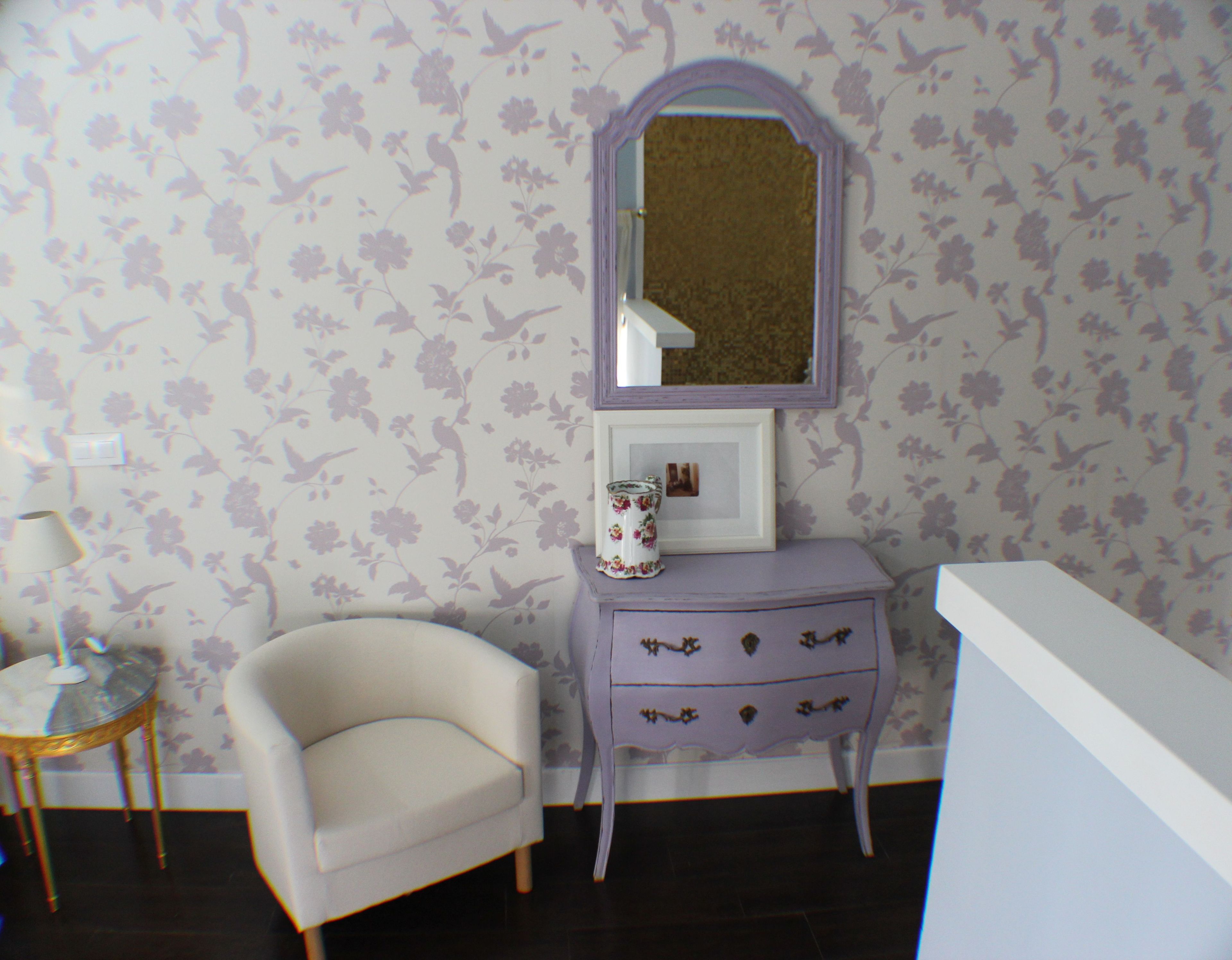 Una habitación con papel tapiz floral, un tocador con espejo, una mesita con lámpara y una silla.