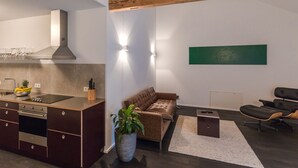 Eine moderne Küche mit Einbauofen, ein Essbereich mit braunem Ledersofa und ein minimalistisches Wohnzimmer mit dunkelbraunem Ledersessel und einem grünen abstrakten Gemälde an der Wand.