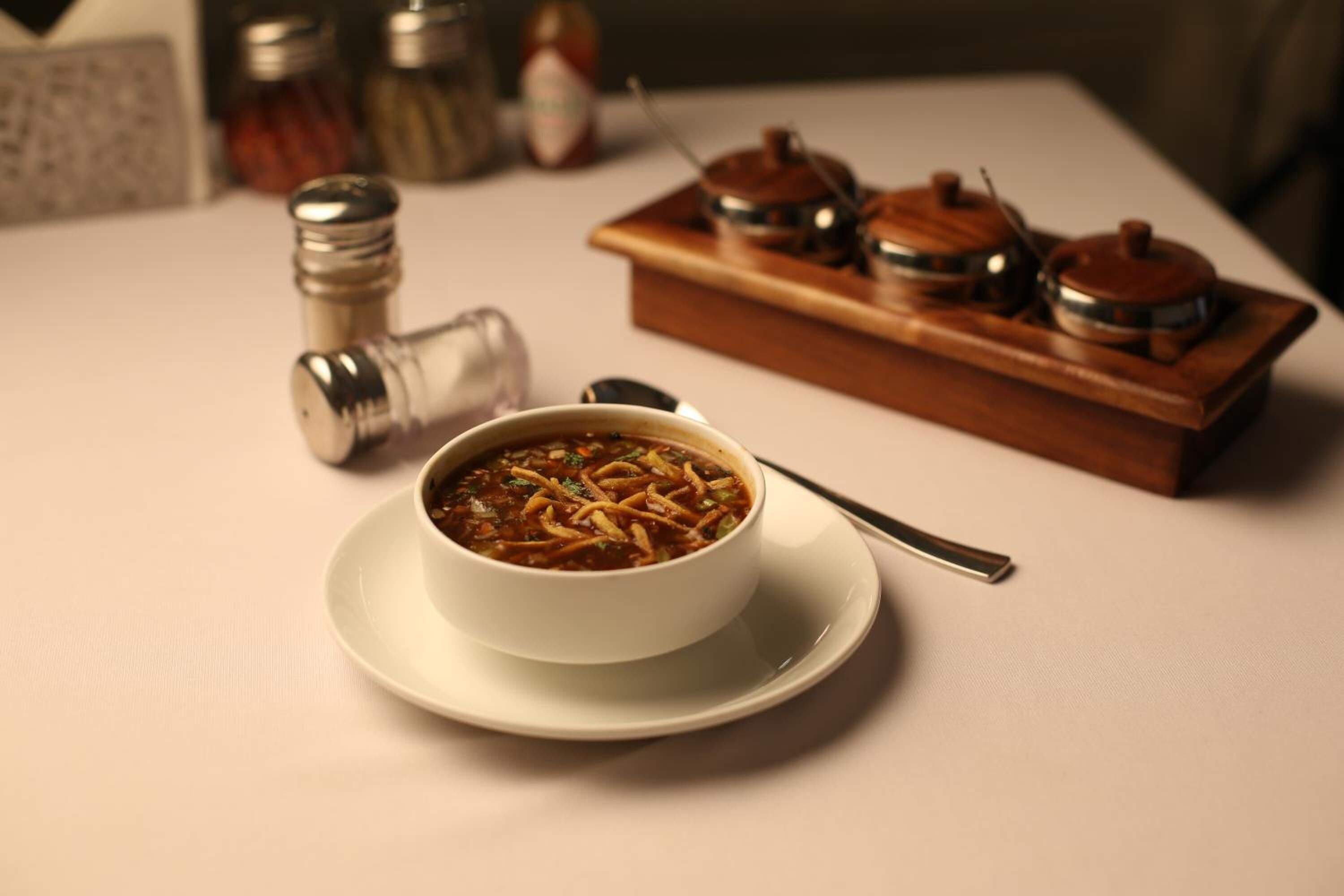A bowl of soup on a white plate with a spoon, a wooden tray with two containers, and salt and pepper shakers on a table.